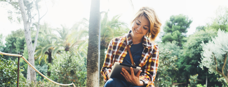 Hipster person holding in hands digital tablet with screen, young girl smile reading on computer on background nature park palm landscape flare, mock up technology blur, female hands tourist using gadgetの写真素材