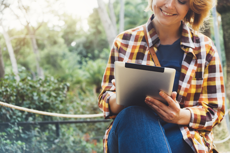 Hipster person holding in hands digital tablet with screen, young girl smile reading on computer on background nature park palm landscape flare, mock up technology blur, female hands tourist using gadgetの写真素材