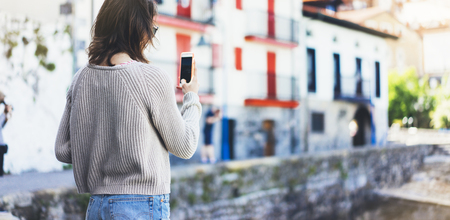 Girl traveler making photo screen smart phone on background old color house in summer sun atmospheric city, young hipster tourist using in female hands mobile phone, mock up flare street, content lifestyleの写真素材