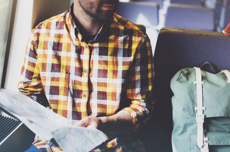Enjoying travel. Young hipster smile man with backpack traveling by train sitting near the window holding in hand and looking map. Tourist in summer shirt planing route of railway, railroad transport conceptの写真素材