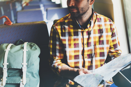 Enjoying travel. Young hipster smile man with backpack traveling by train sitting near the window holding in hand and looking map. Tourist in summer shirt planing route of railway, railroad transport conceptの写真素材