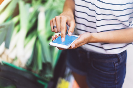 Young woman shopping healthy food in supermarket blur background. Female hands buy products and using mobile smart phone in store. Hipster at grocery holding basket. Person comparing price of produceの写真素材