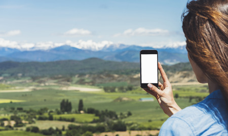 Hipster photograph on smart phone gadget mobile, mock up of blank screen. Girl traveler hold in hand mobile on background mountain landscape horizon. Tourist look map on adventure trip, lifestyle conceptの写真素材
