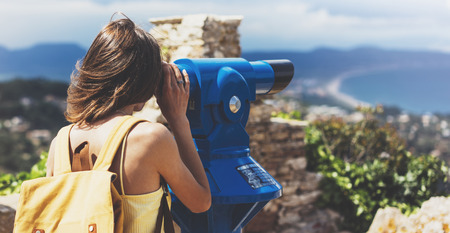 Hipster tourist look observant binoculars telescope on panoramic view, lifestyle concept trip, traveler with backpack on background mountain and blue sea landscape horizon, young girl hiker pointing handsの写真素材