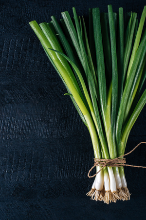 Bunch of green young scallions with roots on a black dark background of the old wooden boards vintage top view, agriculture concept, beetroot onion on cooking process on kitchen table, mock up の写真素材