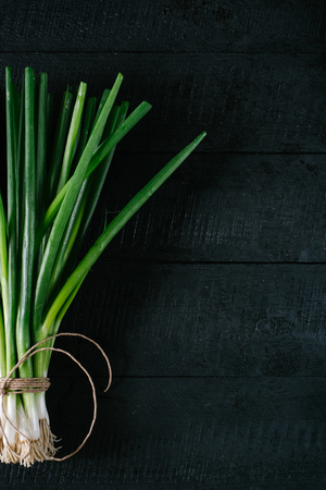 Bunch of green young scallions with roots on a black dark background of the old wooden boards vintage top view, agriculture concept, beetroot onion on cooking process on kitchen table, mock up の写真素材