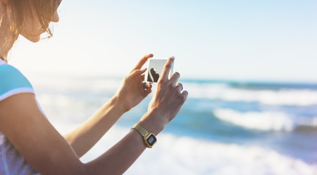 Hipster girl photograph on smart phone gadget in sand coastline, mock up of blank screen. Traveler...