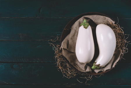 Two white eggplant on textiles with dry grass in a round bowl on wooden mock up, vintage black space background of old dark boards top view, vegan food on table, mock up dieting concept, healthy herbの写真素材