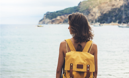 Back view hipster girl with backpack in sand coastline on nature landscape, mock up. Traveler on background beach seascape and horizon mountain. Tourist look on blue sun ocean, summer relax lifestyleの写真素材