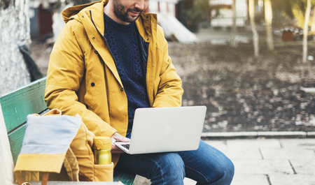 Hipster with yellow backpack, jacket, drink coffee of thermo cup, freelance using computer open laptop in autumn street outdoor, tourist man typing on keyboard, traveler connect wifi internet, process in workspaceの写真素材
