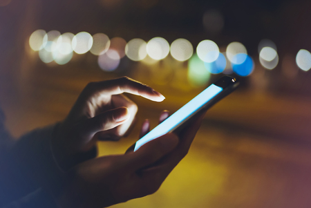Woman pointing finger on screen smartphone on background illumination bokeh light in night atmospheric city, hipster using in hands mobile phone closeup, mockup glitter street in Barcelona, online wifi internet conceptの写真素材