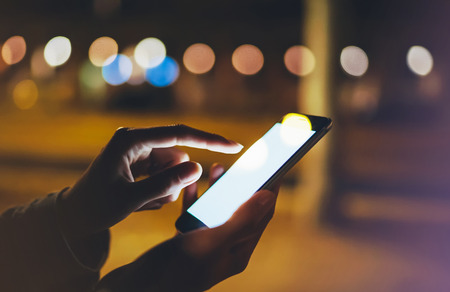 Woman pointing finger on screen smartphone on background illumination bokeh light in night atmospheric city, hipster using in hands mobile phone closeup, mockup glitter street in Barcelona, online wifi internet conceptの写真素材
