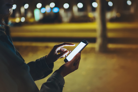 Woman pointing finger on screen smartphone on background illumination bokeh light in night atmospheric city, hipster using in hands mobile phone closeup, mockup glitter street in Barcelona, online wifi internet conceptの写真素材