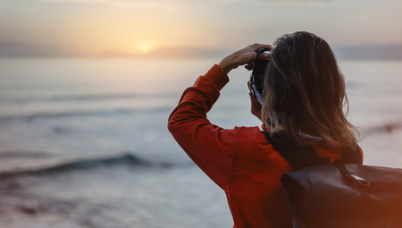 Hipster hiker tourist with backpack taking photo of amazing seascape sunset on camera on background...