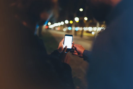 People together pointing finger on screen smartphone on background bokeh light in night atmospheric city, group adult hipsters friends using in hands modern mobile phone closeup, street online wi-fi internet conceptの写真素材