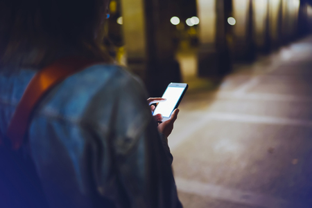 Woman pointing finger on blank screen smartphone on background bokeh light in night atmospheric city, hipster using in hands clean gadget mobile phone closeup, mockup street, online wifi internet conceptの写真素材