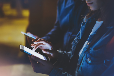 Group adult hipsters using in hands mobile phone closeup, online wi-fi internet concept in street, bloggers friends together pointing finger on screen smartphone on background bokeh light in night atmospheric cityの写真素材