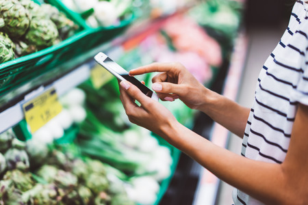 Young woman shopping purchase healthy food in supermarket blur background. Close up view girl blogger buy products using smartphone in store. Hipster at grocery using smartphone. Person comparing priceの写真素材