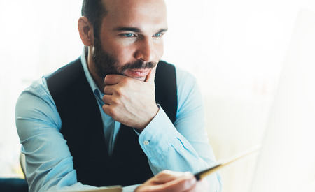 Bearded young businessman working on modern office at night. Consultant man thinking looking in monitor computer. Manager typing on keyboard in coworking workplace, startup project concept in studioの写真素材