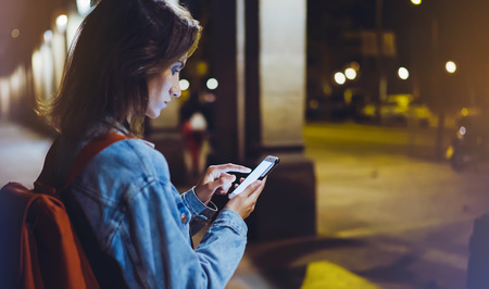 Woman with backpack pointing finger on blank screen smartphone on background bokeh light in night atmospheric city, blogger hipster using in hands gadget mobile phone, mockup street, online wifi internet conceptの写真素材