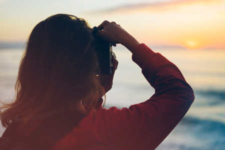 Hipster hiker tourist with backpack taking photo of amazing seascape sunset on camera on background blue sea, photographer enjoying ocean horizon, blurred panoramic sunrise blank mockup, traveler relax holiday concept, sunlight view in trip vacation northern spain の写真素材
