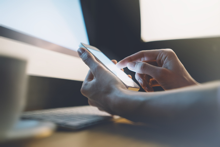 Closeup of a man pointing finger to smartphone with a blank screen monitor in the hands, working behind a computer keyboard and blue monitor on wooden table with flare and blur against a background of bokeh light in the office horizontalの写真素材