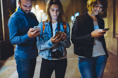 Group adult hipsters using in hands mobile phone closeup, street online wi-fi internet concept, bloggers friends together pointing finger on screen smartphone on background bokeh light in night atmospheric cityの写真素材