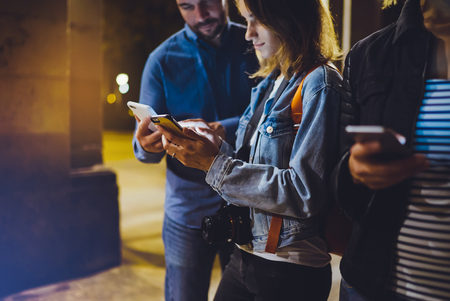 Group adult hipsters using in hands mobile phone closeup, street online wi-fi internet concept, bloggers friends together pointing finger on screen smartphone on background bokeh light in night atmospheric cityの写真素材