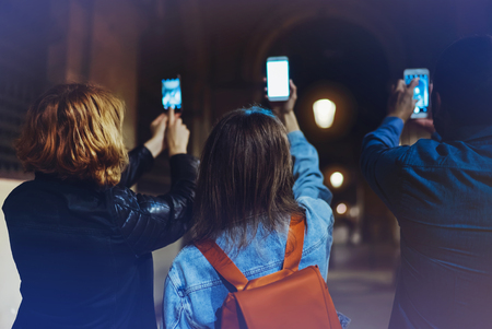 Group adult hipsters using in hands mobile phone closeup, street online wi-fi internet concept, bloggers friends together pointing finger on screen smartphone on background bokeh light in night atmospheric cityの写真素材