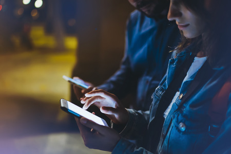 Group adult hipsters using in hands mobile phone closeup, online wi-fi internet concept in street, bloggers friends together pointing finger on screen smartphone on background bokeh light in night atmospheric cityの写真素材