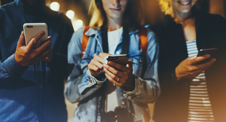 Group adult hipsters friends using in hands mobile phone closeup, street online wi-fi internet concept, bloggers together pointing finger on screen smartphone on background bokeh light in night atmospheric cityの写真素材