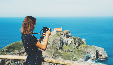 Tourist traveler photographer making pictures sea scape on photo camera on background ocean gaztelugatxe steps on Spain, hipster girl looking on nature horizon, relax holiday, blank space blue waves viewの写真素材