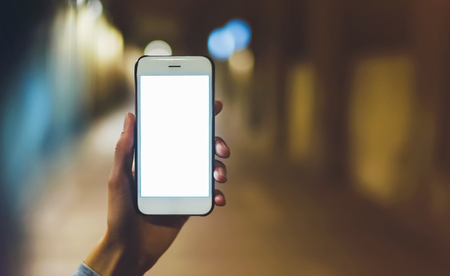 hipster using in hands clean gadget mobile phone closeup, blogger pointing finger on blank screen smartphone on background bokeh light in night atmospheric city, mockup street, online wifi internet conceptの写真素材