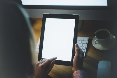 Girl using a tablet with a clean blank screen display in business office atmosphere on the keyboard and cup of coffee on the wooden table background, digital gadget in female hands in the night workspace atmosphereの写真素材