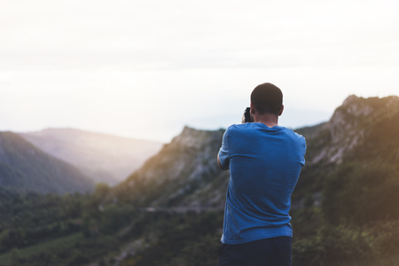 Hipster man enjoying sunset on peak of foggy mountain, tourist traveler taking pictures of amazing landscape on digital cameraの写真素材