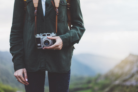 Hipster girl with backpack enjoying sunset on peak of foggy mountain, tourist traveler photographer taking pictures of amazing landscape on vintage photo cameraの写真素材