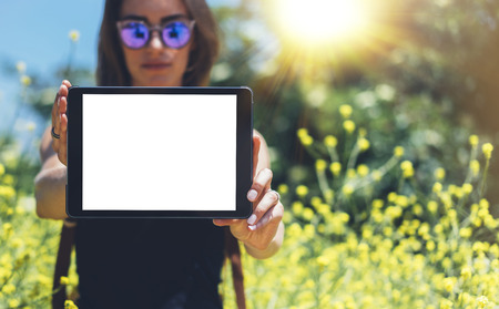 Hipster holding in hands tablet computer. Girl traveler with sunglasses using gadget on sun flare and yellow flowers backgroundの写真素材