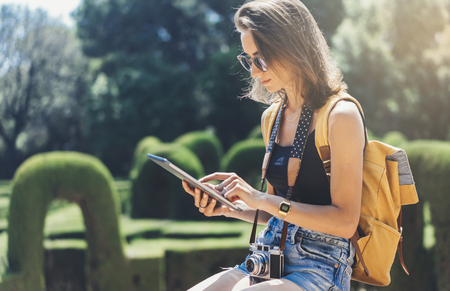 Hipster girl with backpack, sunglasses and vintage photo camera using tablet computer, copy space of blank empty mockup, view tourist hands holding gadget and texting message on device travel on background sun flareの写真素材