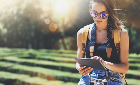 Hipster girl with backpack, sunglasses and vintage photo camera using tablet computer, copy space of blank empty mockup, view tourist hands holding gadget and texting message on device travel on background sun flareの写真素材