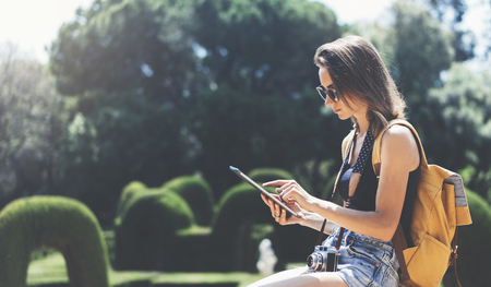 Hipster young girl with bright backpack and vintage camera using tablet or holding gadget, planning travel plan. View tourist traveler with sunglasses on background Parc Laberint Barcelona. Mockup for text messageの写真素材