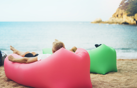 young hipster relaxing on coastline beach on inflatable lazy air pouffe sofa, person tourist enjoy sunny day on background coast sea waves, calm concept mockup, seascape perspective vacation, summer seashoreの写真素材