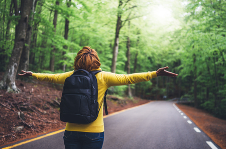 tourist traveler with backpack standing with raised hands, girl hiker view from back looking into road at forest with arms outstretched and enjoying the breath of fresh clean air in trip, relax holiday conceptの写真素材
