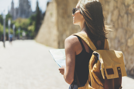 Hipster young girl with sunglasses and bright backpack using and looking at map. Said view tourist traveler enjoying on background panoramic city. Person visiting the sights Barcelona in holiday. Mockupの写真素材