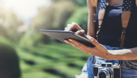 Hipster girl with backpack and vintage photo camera using tablet computer close up, copy space of blank empty mockup, view tourist hands holding gadget and texting message on device travel on background sun flareの写真素材