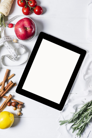 Tablet with a measuring tape, rosemary, nuts, parsley root, tomatoes, cinnamon and red apples on a white wooden background top view verticalの写真素材