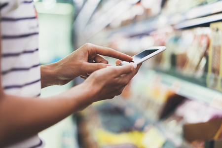 Woman shopping healthy food in supermarket background. Close up view girl buy products using digital gadget in store. Hipster at grocery using smartphone. Person comparing the price of produceの写真素材