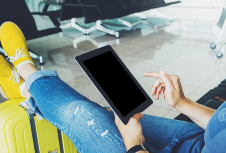 Young hipster girl sitting at airport in yellow boot on suitcase traveling in Europe, female hands texting message on gadget in terminal area hall, summer journey trip concept, mockup blank screen tabletの写真素材