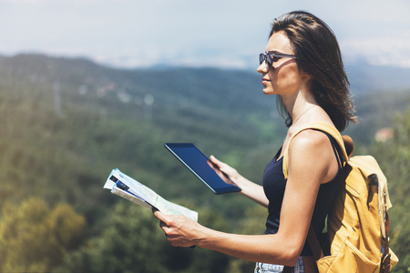 Hipster young girl with backpack enjoying panoramic mountain sea, using map and looking at horizon. Tourist traveler texting on tablet computer on background landscape Barcelona view mockup, sunの写真素材