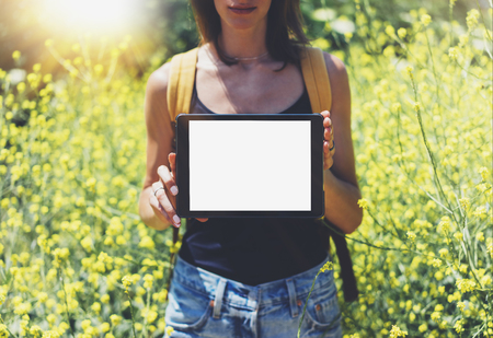 Nature view hipster holding in hands tablet computer. Girl traveler using gadget on sun flare and yellow flowers background, mockup of blank empty screen copy space for text, female photographerの写真素材