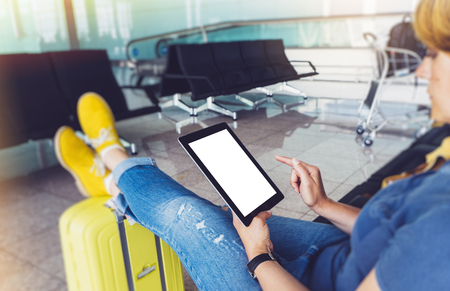 Young hipster girl at airport in yellow boot on suitcase waiting air flight, female hands holding computer in terminal departure lounge gate, traveler trip concept, mock up of blank screen tabletの写真素材